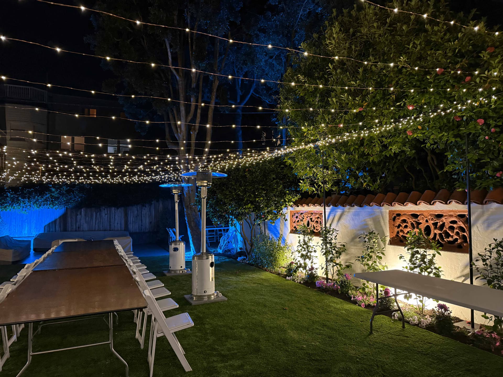 Backyard event at night with a multi-layered warm white string light canopy, blue uplighting, and set dining tables
