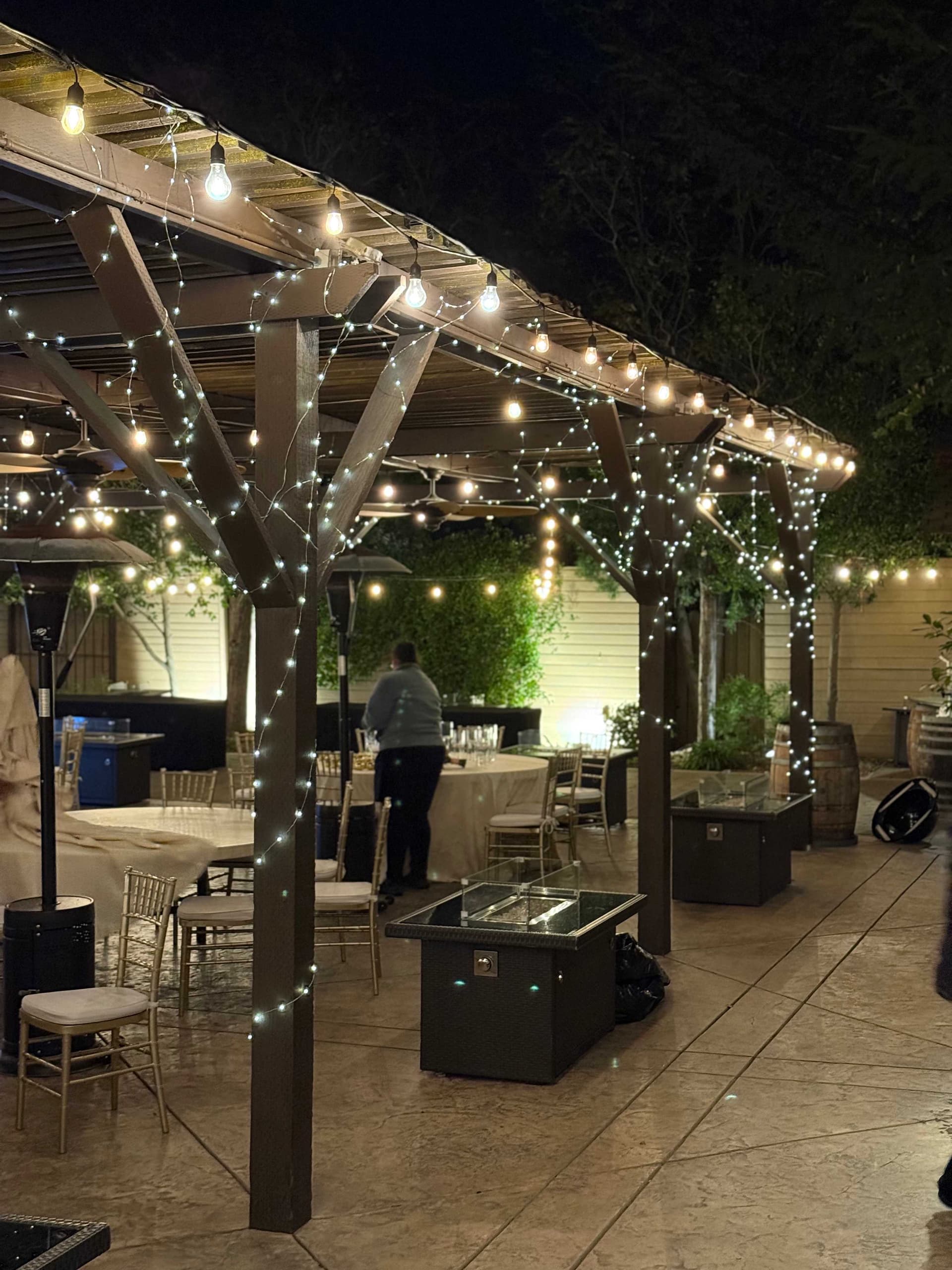 Edison string lights and fairy lights wrapped around a dark-wood pergola at a reception
