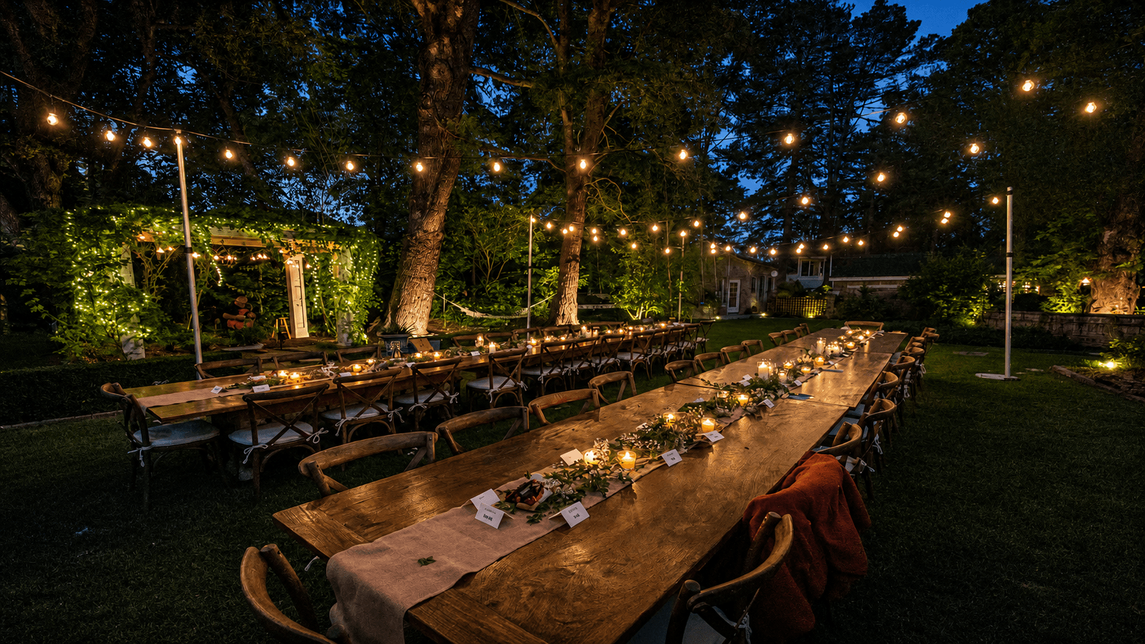 Twilight Bay Area estate backyard dinner with long farm tables, candle and eucalyptus runners, and a warm bistro string light canopy strung between trees over the lawn